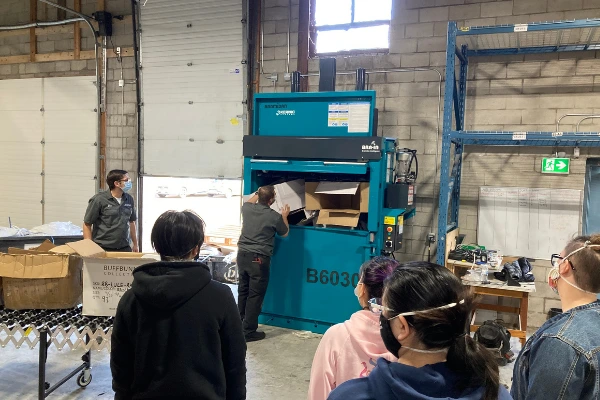 two men loading teal baler with cardboard while others look on