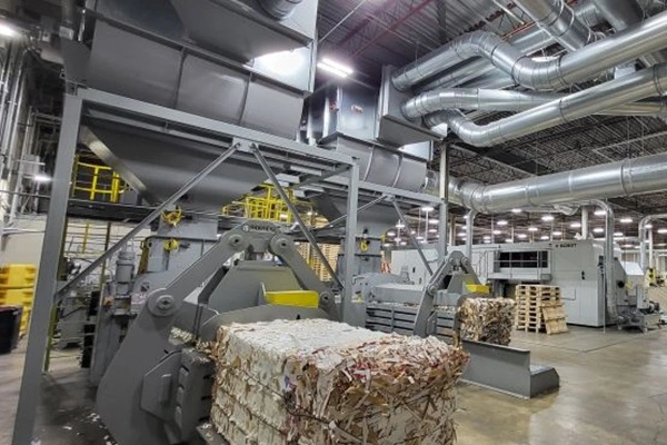 Industrial recycling plant scene with a large baler and conveyor system; foreground shows a compressed paper bale and overhead ductwork.