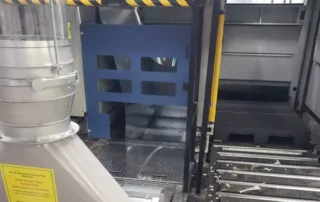 Industrial work area with blue safety gate, grated metal floor, and yellow/black caution tape overhead; large duct/tube on left and metal stairs on right.