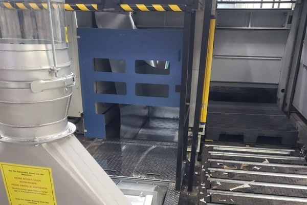 Industrial work area with blue safety gate, grated metal floor, and yellow/black caution tape overhead; large duct/tube on left and metal stairs on right.