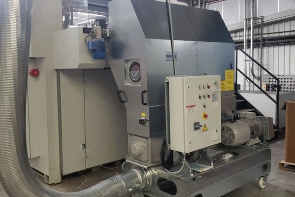 Industrial processing equipment in a factory: a large metal machine with a control panel, ducts, and a motor on a wheeled base.