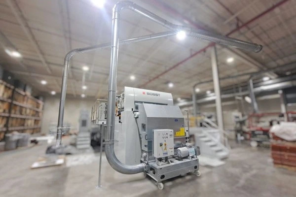 Industrial processing machine in a spacious warehouse, connected to metal ductwork with overhead lighting.