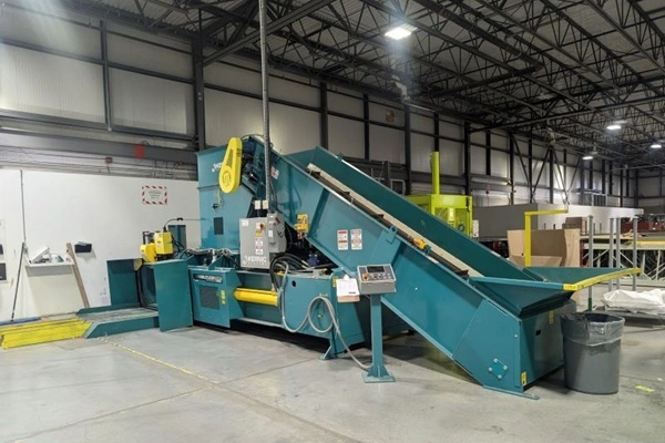 Teal industrial processing machine with a sloped conveyor feeding into a large hopper, located in a warehouse with metal beams and overhead lighting.