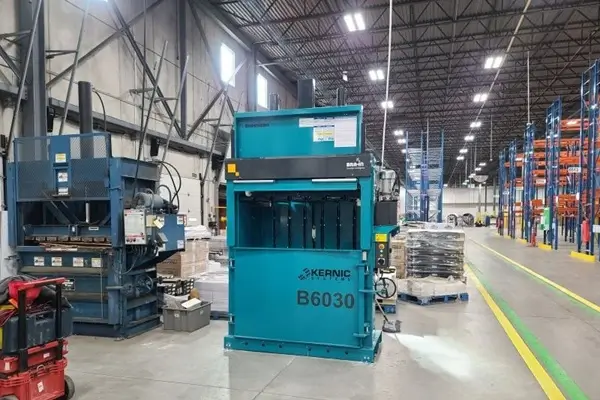 Blue industrial vertical baler labeled B6030 in a warehouse, with pallets and shelving visible in the background.