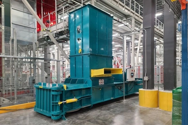 Large teal industrial baler/shredder in a factory, with yellow safety rails and metal framework surrounding it.