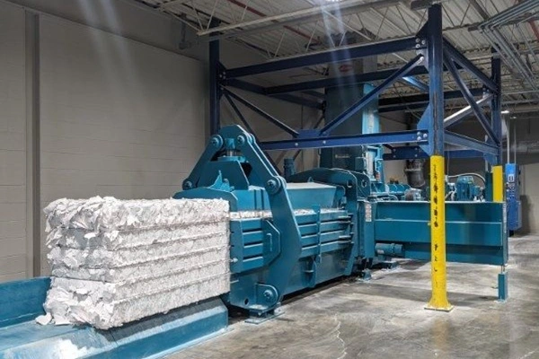 Blue industrial baler or compactor in a warehouse, with a bale of white packing material fed into the machine