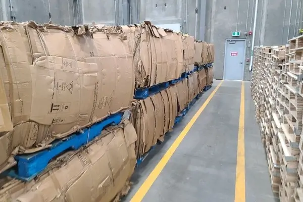 Warehouse aisle with tall stacks of flattened cardboard bales on blue pallets along the left; wooden pallets on the right and a gray door at the far end.