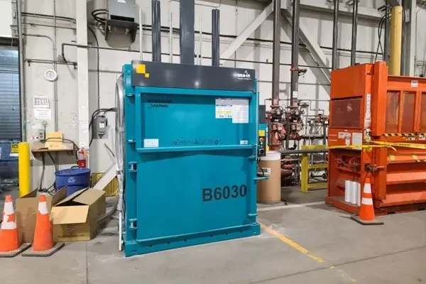 Blue industrial vertical baler unit labeled B6030 in a factory setting, with an orange waste container and safety cones nearby.