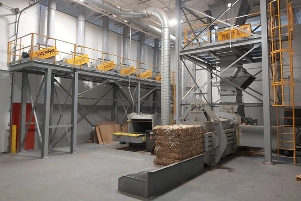 Industrial processing facility with a tall metal framework, yellow safety rails, overhead ducts, and a large baling/compression machine feeding a bundled material bale.