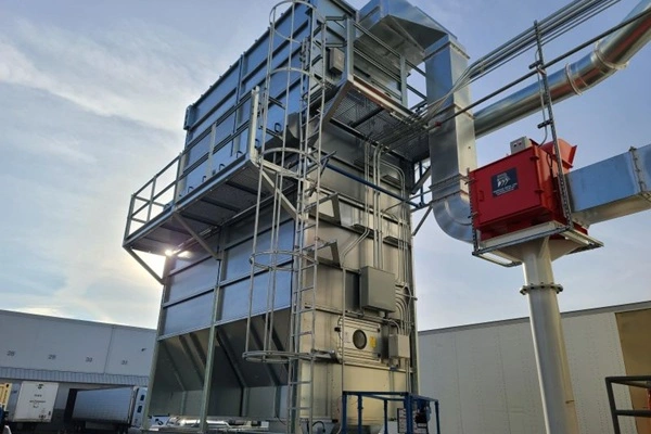 Tall metal processing tower with stairs and platforms, connected to large ductwork and a red control box, outside a warehouse.