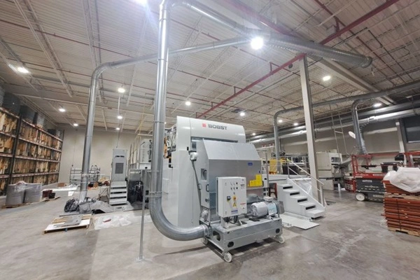 Large industrial processing machine in a spacious factory, with overhead ducts, stairs, and storage shelves nearby.