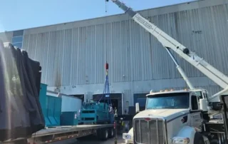 Crane lifting a large piece of industrial equipment from a flatbed trailer beside a warehouse.