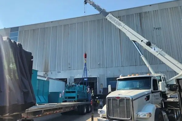 Crane lifting a large piece of industrial equipment from a flatbed trailer beside a warehouse.