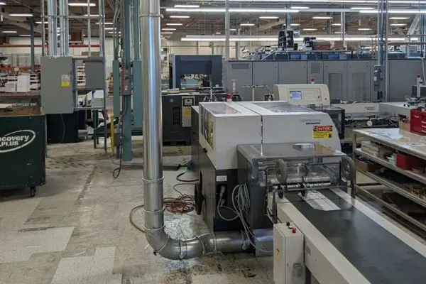 Industrial machinery in a factory: a large white processing unit with a conveyor belt and attached ductwork amid other equipment and workstations.