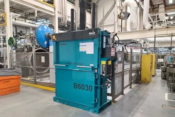 Industrial metal stamping press in teal with 'B6030' label, surrounded by safety fencing in a manufacturing plant, with overhead piping and equipment nearby.