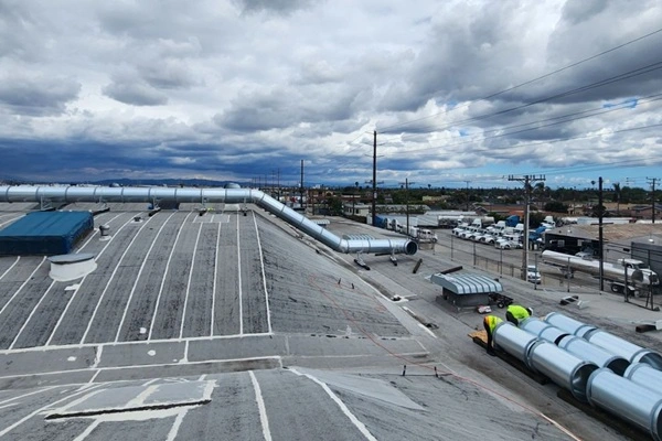 Industrial rooftop with large silver HVAC ducts and a worker in a high-visibility vest against a cloudy sky beside curved piping.