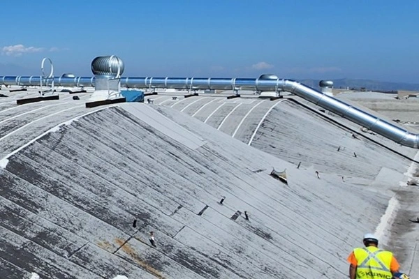 Industrial rooftop with large silver ventilation ducts and workers in safety vests inspecting the slope in clear weather.