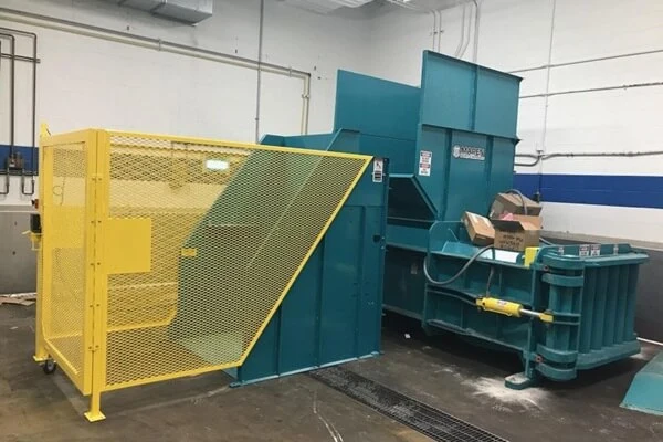 Industrial baler with yellow safety cage feeding a blue compaction unit; cardboard boxes sit near a green recycling bin in a warehouse.