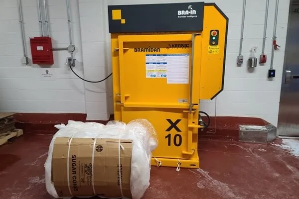 Yellow Bramidan X10 baler in a warehouse, with a large wrapped cardboard bale beside it.