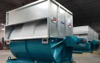 Two large industrial machines with teal lower sections and light gray upper housings on a factory floor, featuring red valve handles and mounted on wooden blocks